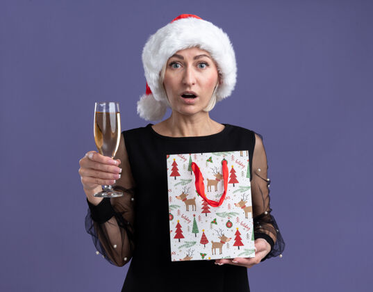金发令人印象深刻的中年金发女人戴着圣诞帽拿着一杯香槟和圣诞礼包隔离在紫色的墙壁上与复制空间香槟杯子女人