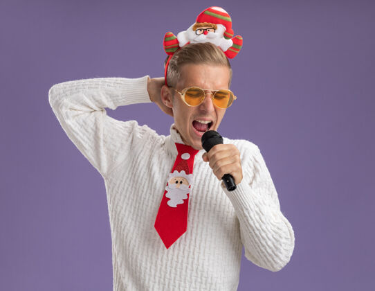 小伙子年轻帅哥戴着圣诞老人的头带和领带戴着眼镜拿着麦克风手放在脑后唱歌闭着眼睛隔离在紫色背景上保管眼镜眼睛