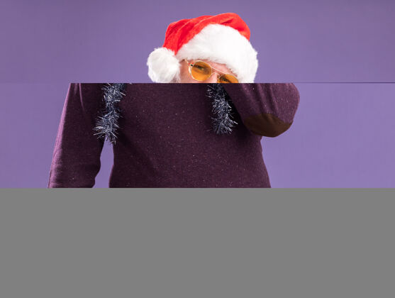 脖子困惑的中年男子戴着圣诞帽 脖子上戴着金箔花环 戴着眼镜 手放在脸上 隔离在紫色的墙上困惑保管花环