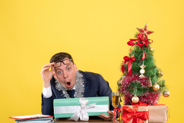 办公室大眼睛男人摘下眼镜坐在圣诞树旁的桌子前 呈现黄色圣诞礼物新郎