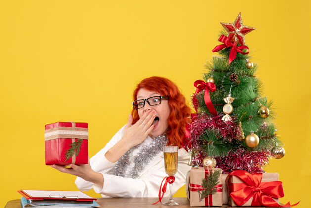 圣诞节前视图：女医生坐在桌子后面 黄色背景上有圣诞礼物快乐情感微笑
