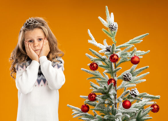 穿关心的小女孩站在圣诞树旁 戴着头饰 脖子上戴着花环 手放在脸颊上 隔离在橙色的墙上关心头饰脸颊