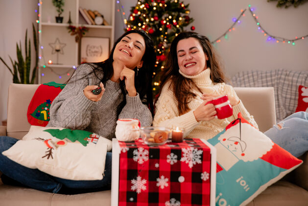 微笑笑容可掬的年轻女孩拿着电视遥控器和杯子坐在扶手椅上 在家里享受圣诞节时光享受坐着圣诞节