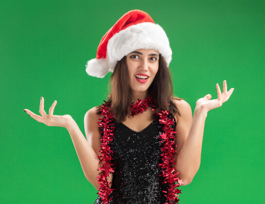 帽子微笑的年轻漂亮女孩戴着圣诞帽 脖子上戴着花环 双手分开放在绿色的墙上圣诞节蔓延脖子