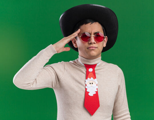 帽子自信的年轻人戴着帽子 打着圣诞领带 戴着眼镜 在绿色的墙上展示着敬礼的姿势展示敬礼圣诞节