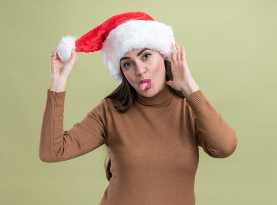 帽子快乐的歪头年轻漂亮的女孩戴着圣诞帽在橄榄绿的墙上露出孤立的舌头快乐舌头圣诞节