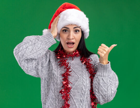 关心关心的年轻白人女孩戴着圣诞帽 脖子上戴着金箔花环 手放在头上 指着绿色墙壁上与世隔绝的一面侧面周围穿着
