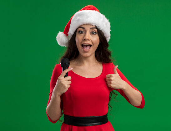 女孩令人印象深刻的年轻漂亮女孩戴着圣诞帽拿着麦克风看着相机显示拇指上孤立的绿色背景麦克风拇指漂亮