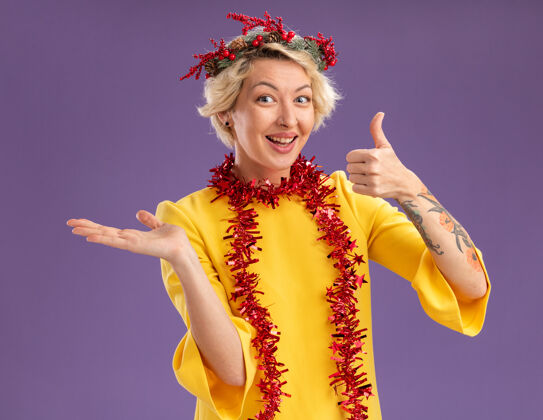 金发快乐的年轻金发女人戴着圣诞花环 脖子上戴着金箔花环 看起来空手而起 拇指孤立地站在紫色的墙上头空穿着