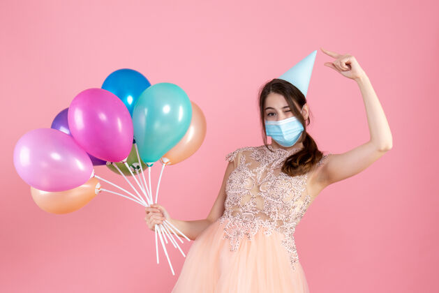 举行戴着医用面具的派对女孩指着她的派对帽 手里拿着粉色的彩色气球气球派对庆典