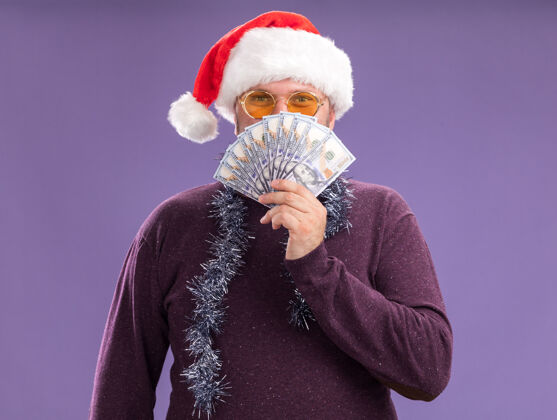 钱高兴的中年男子戴着圣诞帽 脖子上戴着金箔花环 戴着眼镜 从后面把钱隔在紫色的墙上戴着高兴男人