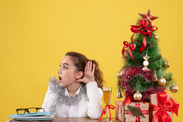 健康正面图：女医生坐在桌子后面 听着黄色背景下的圣诞树和礼品盒人新郎医生
