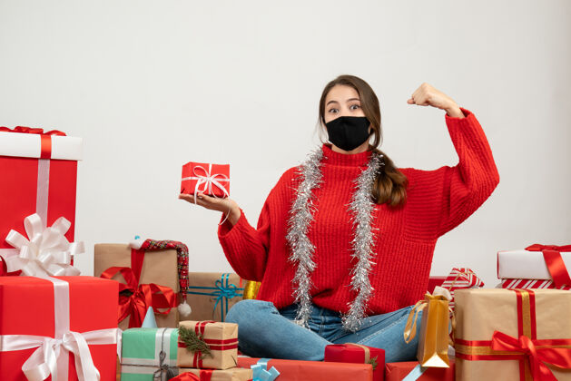 圣诞节穿着红色毛衣 戴着黑色面具的年轻女孩拿着礼物展示她的力量坐在白色的礼物旁边黑色坐着力量