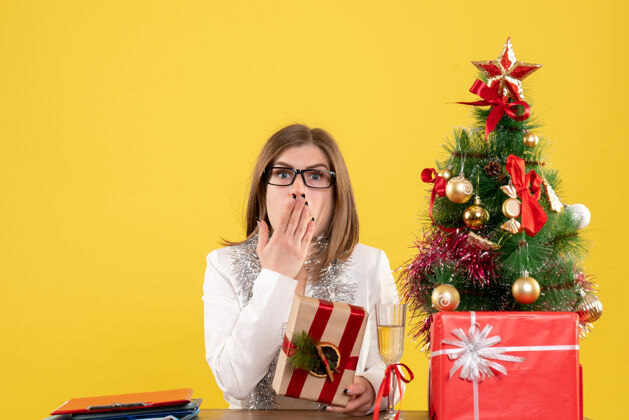 新正面图：女医生坐在桌子前 拿着黄色背景的礼物 还有圣诞树和礼品盒现在生活医生