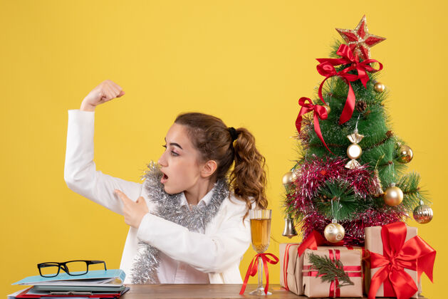 人前视图：女医生坐在桌子后面 在黄色背景上弯曲着 带着圣诞树和礼品盒屈曲快乐成人