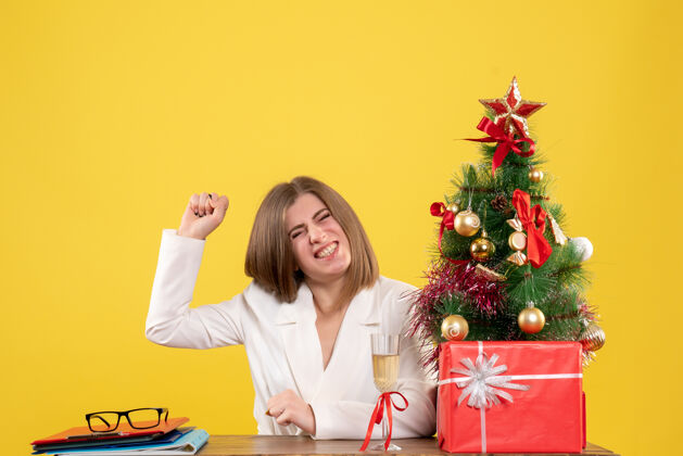 背景正面图：女医生坐在黄色背景的桌子前 放着圣诞树和礼品盒女医生情绪医院
