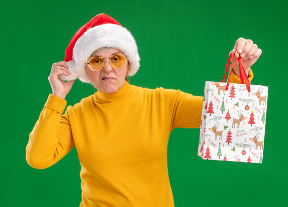 包戴着太阳眼镜 戴着圣诞帽 拿着纸质礼品袋的恼怒老妇人被隔离在绿色背景上 还有复印空间快乐帽子烦恼