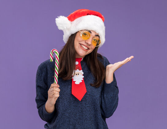 年戴着太阳眼镜 戴着圣诞帽 系着圣诞领带 面带微笑的年轻白人女孩手持糖果手杖 在紫色背景上保持手张开 并留有复印空间女孩帽子微笑