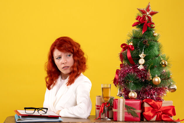 安排女工人坐在桌子后面 手里拿着圣诞礼物和黄色的圣诞树坐着情绪礼物