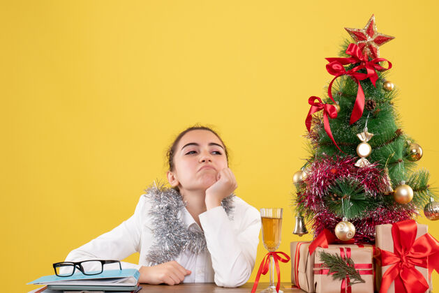 树前视图：女医生坐在桌子后面 拿着圣诞礼物 黄色背景下的树感觉无聊前面坐圣诞节