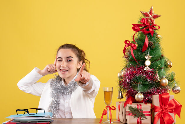人前视图：女医生坐在桌子后面 拿着圣诞礼物和黄色背景上的树前面礼物女