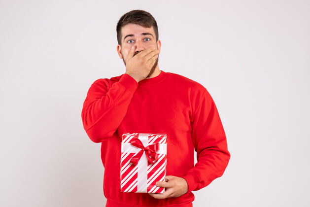 礼物一个穿着红衫的年轻人在白色的墙上举着圣诞礼物的正视图年轻的男人快乐人