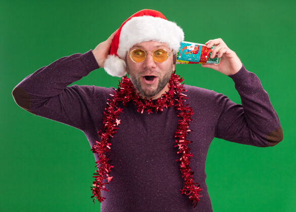 看印象深刻的中年男子戴着圣诞帽 脖子上戴着金箔花环 戴着眼镜 耳边拿着塑料圣诞杯 听着保密的手帽子中年举行