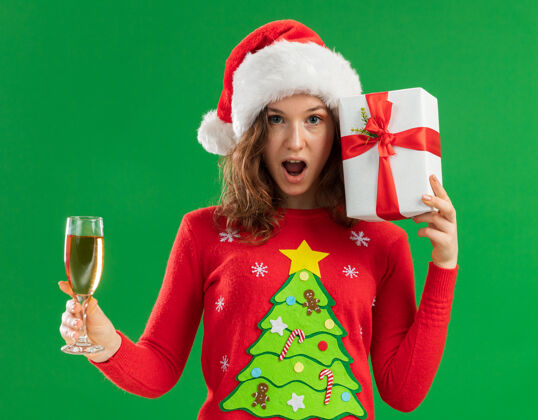 毛衣一位身穿红色圣诞毛衣 戴圣诞帽 手持香槟酒杯的年轻女士站在绿色的背景下 兴高采烈地看着镜头拿着年轻人杯子