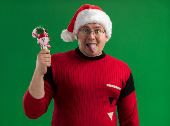 眼镜可爱的成年男子戴着眼镜 戴着圣诞帽 手里拿着糖果手杖 在绿色的背景上露出孤立的舌头看圣诞老人好玩
