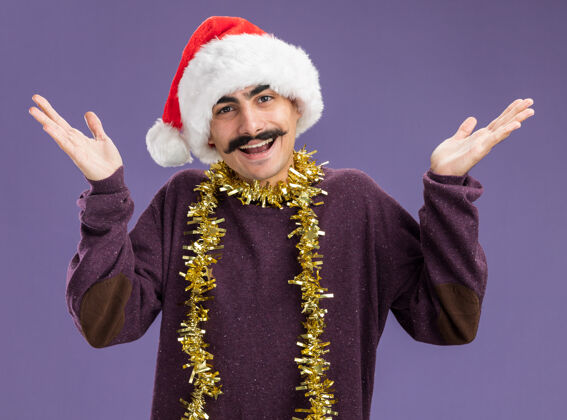 周围年轻的留胡子的男人戴着圣诞老人的帽子 脖子上戴着金属丝 高兴而兴奋地看着相机 双臂举过紫色的背景圣诞老人站立脖子