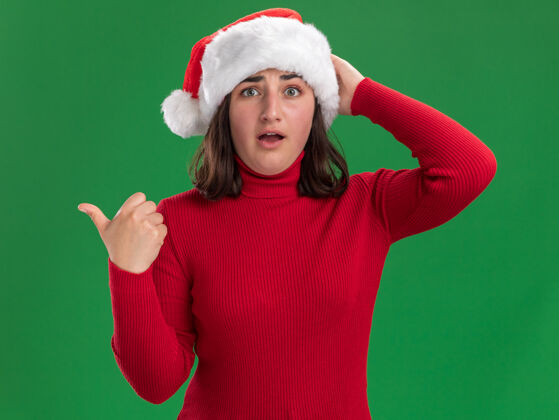 圣诞老人穿着红毛衣 戴着圣诞帽的小女孩站在绿色的墙上 头上的手被弄糊涂了站着年轻圣诞节