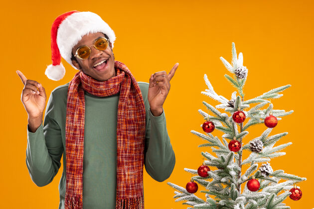 高兴地戴着圣诞帽 脖子上围着围巾的非裔美国人站在橙色墙上的圣诞树旁 开心地微笑着站着男人围巾