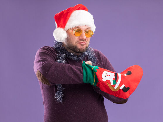 长袜好奇的中年男子戴着圣诞帽 脖子上戴着金属丝的花环 戴着眼镜 手里拿着圣诞长袜 看着里面的手 孤零零地站在紫色的墙上周围男人放
