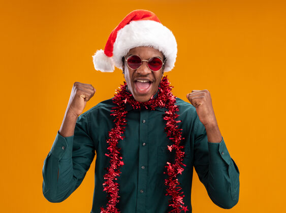 男人戴着圣诞帽 戴着花环 戴着眼镜的非裔美国人站在橙色的墙上 高兴而兴奋地握紧拳头圣诞老人花环握紧