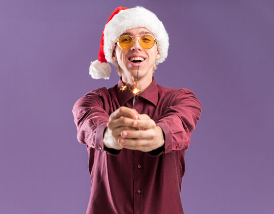 男人快乐的年轻金发男子戴着圣诞帽和眼镜伸出节日火花孤立在紫色墙上的复制空间快乐穿着年轻