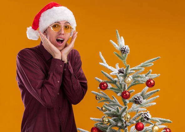 金发兴奋的年轻金发男子戴着圣诞帽和眼镜站在装饰过的圣诞树旁 手靠近橙色墙上的隔离脸穿着兴奋保持