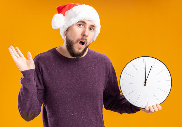 肩膀一个穿着紫色毛衣 戴着圣诞老人帽子的年轻人拿着挂钟 困惑而焦急地耸耸肩站在橙色的墙上困惑帽子非常