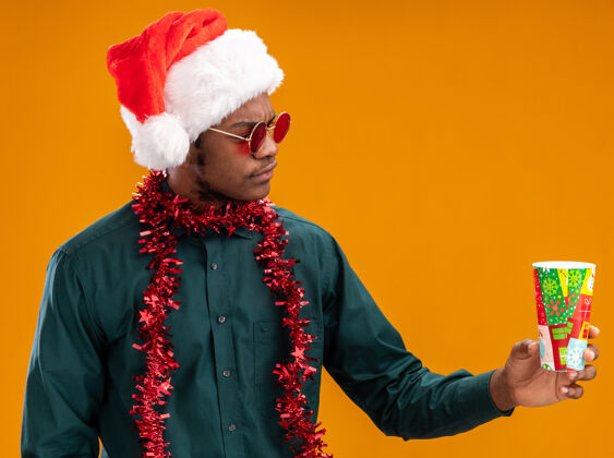 穿着一个戴着圣诞帽 戴着花环 戴着墨镜 拿着五颜六色纸杯的非裔美国人站在橙色的墙上严肃地看着它圣诞老人五颜六色杯子