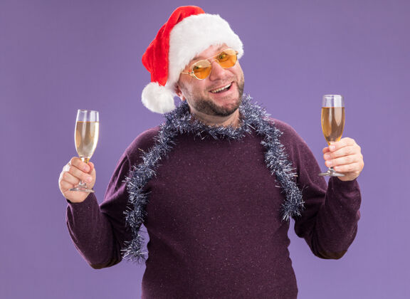 花环快乐的中年男子戴着圣诞帽 脖子上戴着金箔花环 戴着一副眼镜 手里拿着两杯香槟 隔离在紫色的墙上圣诞老人帽子脖子