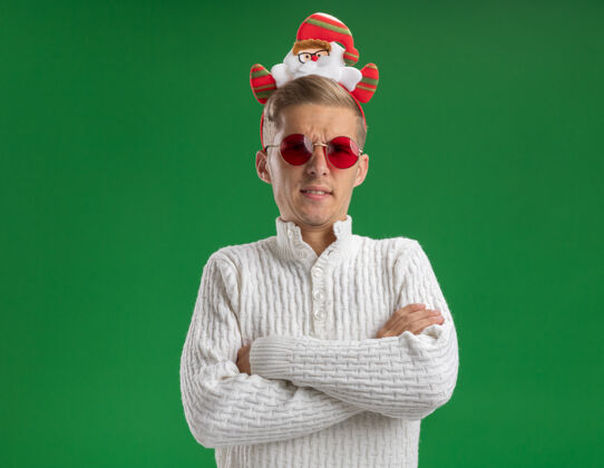 年轻愁眉苦脸的年轻帅哥戴着圣诞老人的头带 戴着眼镜 站着封闭的姿势 隔离在绿色的墙上 留着复制空间头带圣诞老人戴