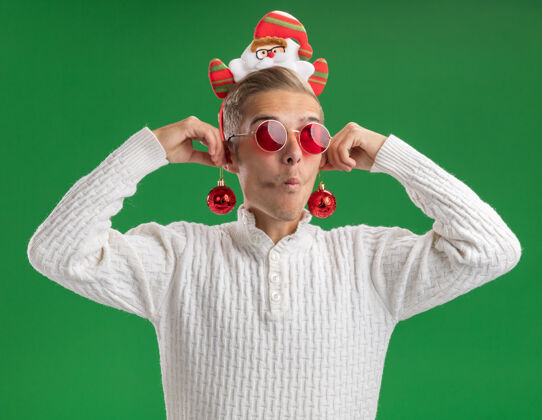 圣诞节给人印象深刻的年轻帅哥戴着圣诞老人的头带戴着眼镜把圣诞饰品挂在耳朵上看着一边 噘着嘴唇隔离在绿色的墙上年轻侧印象深刻