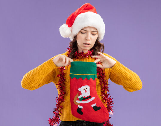 周围无知的年轻斯拉夫女孩戴着圣诞帽 脖子上戴着花环 拿着圣诞长袜 看着紫色背景上的复制空间举行女孩圣诞老人