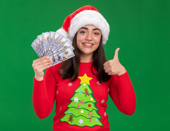 微笑带着圣诞帽的年轻高加索女孩微笑着拿着钱 竖起大拇指 孤立地站在绿色背景上 留着复制空间拇指年轻圣诞老人