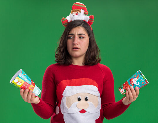 困惑穿着圣诞毛衣戴圣诞帽拿着五颜六色纸杯的年轻女孩困惑地看着他们试图做出选择有疑虑地站在绿色的墙上杯子圣诞节制作