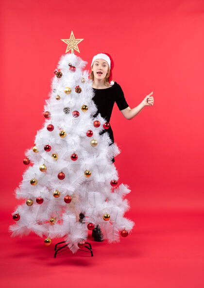 雪人新年的气氛与年轻女子在黑色礼服和圣诞老人的帽子站在白色圣诞树后面圣诞老人圣诞树站着