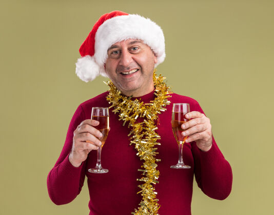 站立快乐快乐的中年男子戴着圣诞老人帽 脖子上戴着金属丝 手里拿着香槟酒杯 站在绿色的背景下微笑着看着相机中间年龄脖子