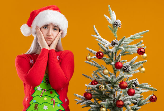 橙色身穿圣诞毛衣 戴圣诞帽的金发女郎站在橙色背景下的圣诞树旁 神情悲伤地看着摄像机 双臂放在脸颊上圣诞快乐圣诞树金发