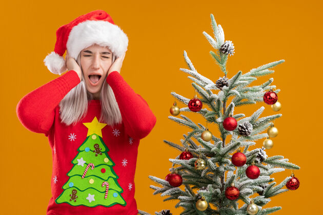 毛衣身穿圣诞毛衣 戴圣诞帽 双手捂着耳朵 表情烦躁的金发女郎站在橙色背景下的圣诞树旁手圣诞快乐快乐