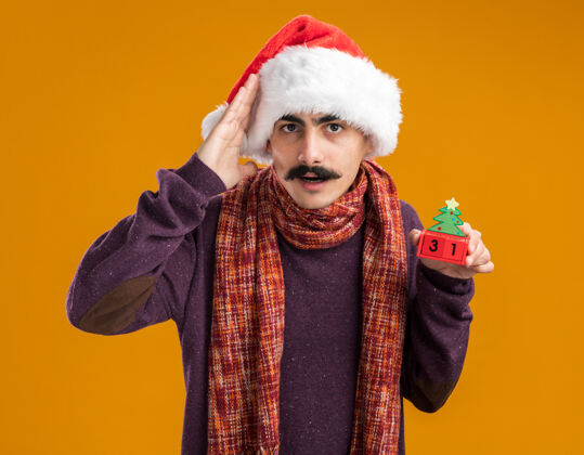 站着一个留着胡子的男人戴着圣诞老人帽 脖子上围着暖和的围巾 手里拿着玩具方块 上面写着“25岁”的日期 看着相机 困惑地站在橙色的背景上 用手捂着头头立方体周围