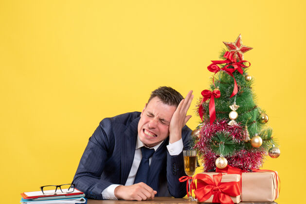 男人焦躁不安的男人闭着眼睛坐在圣诞树旁的桌子前 黄色的墙上挂着礼物礼物圣诞节关门